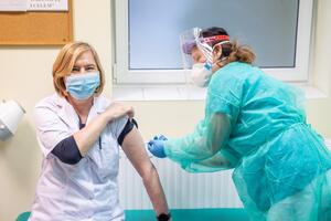 Kobieta o blond włosach siedzi w pomieszczeniu medycznym, jest po lewej stronie zdjęcia. Po prawej - pielęgniarka w zielonym fartuchu i masce ochronnej, która przystępuje do wykonania zastrzyku