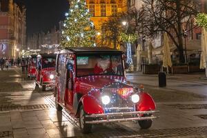 czerwone samochody elektryczne na Długim Targu, przybrane w lampki choinkowe, za kierownicą Mikołaj pokazujący znak ok