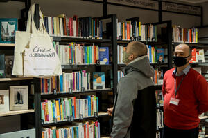 czarne metalowe regały biblioteczne zastawione książkami, z lewej strony na regale wiszą materiałowe torby w kolorze lnu z napisem Chodzę do Biblioteki!. Na torby patrzy mężczyzna w szaro-czarnej bluzie i w zielonej maseczce. Obok stoi pracownik biblioteki w niebieskiej koszuli i czerwonym swetrze, na szyi ma identyfikator, na twarzy czarną maseczkę.