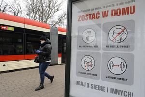 Fragment tramwaju, przechodzi kobieta w maseczc, na pierwszym planie plakat zachęcający do zostania w domu
