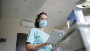 (zdjęcie od dołu): kobieta w maseczce i fartuchu w sali szpitalnej. W dłoniach trzyma kartę medyczną, przed nią fragment łóżka, za nią drzwi, ściana i sufit