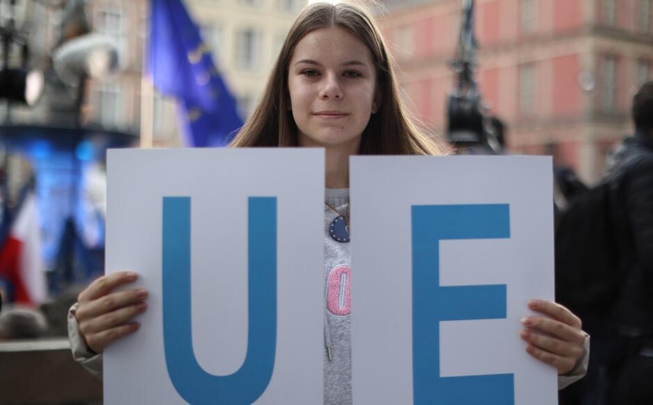 Na zdjęciu widzimy młodą kobietę, która trzyma dwa białe plakaty z literami "U" i "E" w kolorze niebieskim, które razem tworzą skrót "UE", co jest powszechnie używanym skrótem dla Unii Europejskiej. Kobieta ma długie, proste brązowe włosy i jest ubrana w szary top. Stoi na tle zatłoczonego miejsca, prawdopodobnie na jakimś zgromadzeniu lub demonstracji, co sugerują nieostre tło i widoczne flagi. Na jej twarzy maluje się delikatny uśmiech.